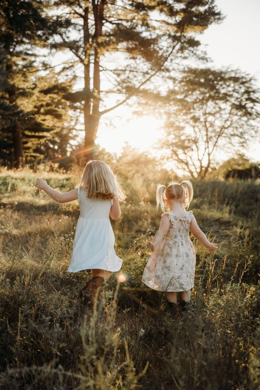 Two young girls in summer dresses running through a sunlit meadow toward golden trees at sunset.