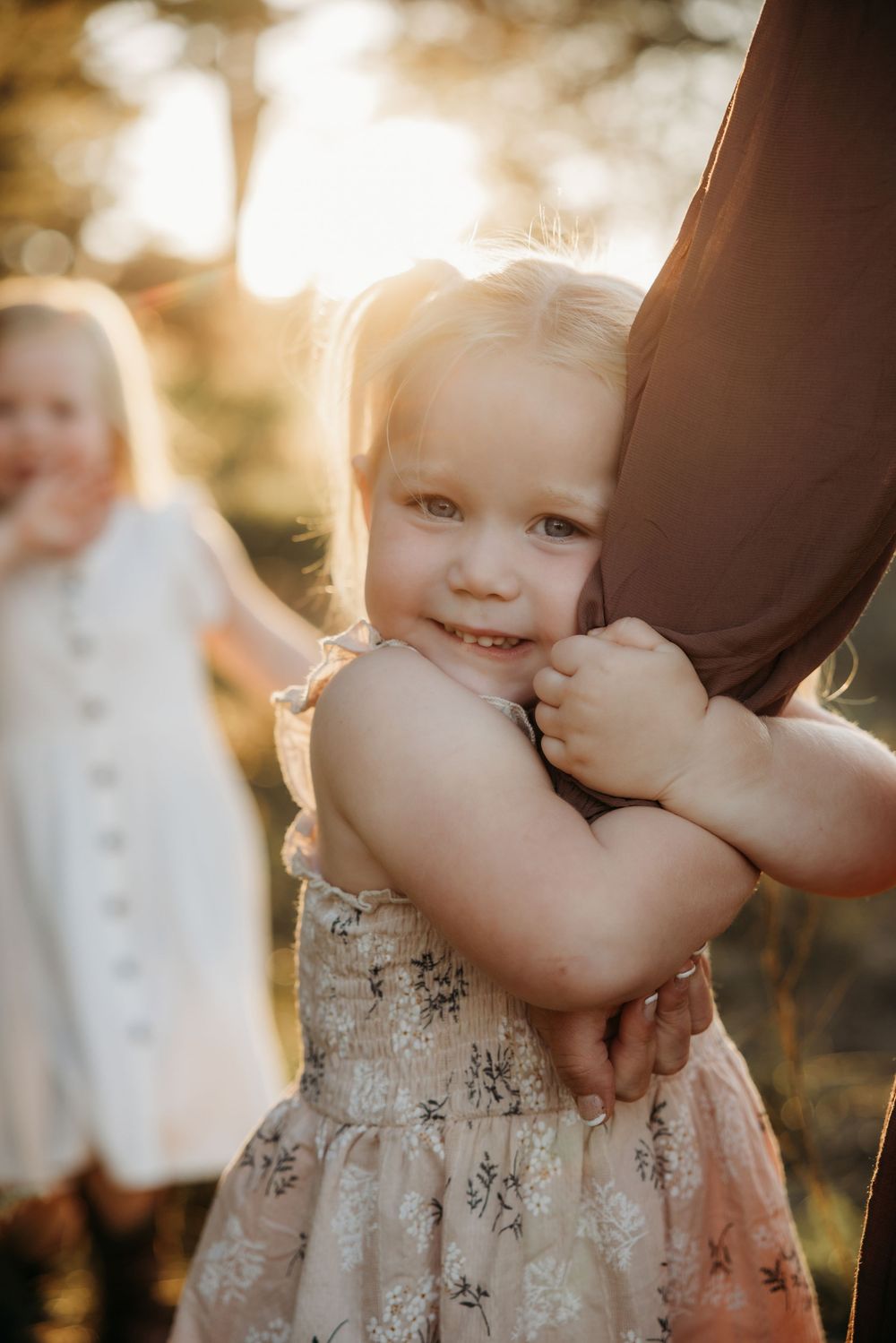 Toddler girl in floral dress hugging an adult's leg outdoors in golden sunset light with another child in background.