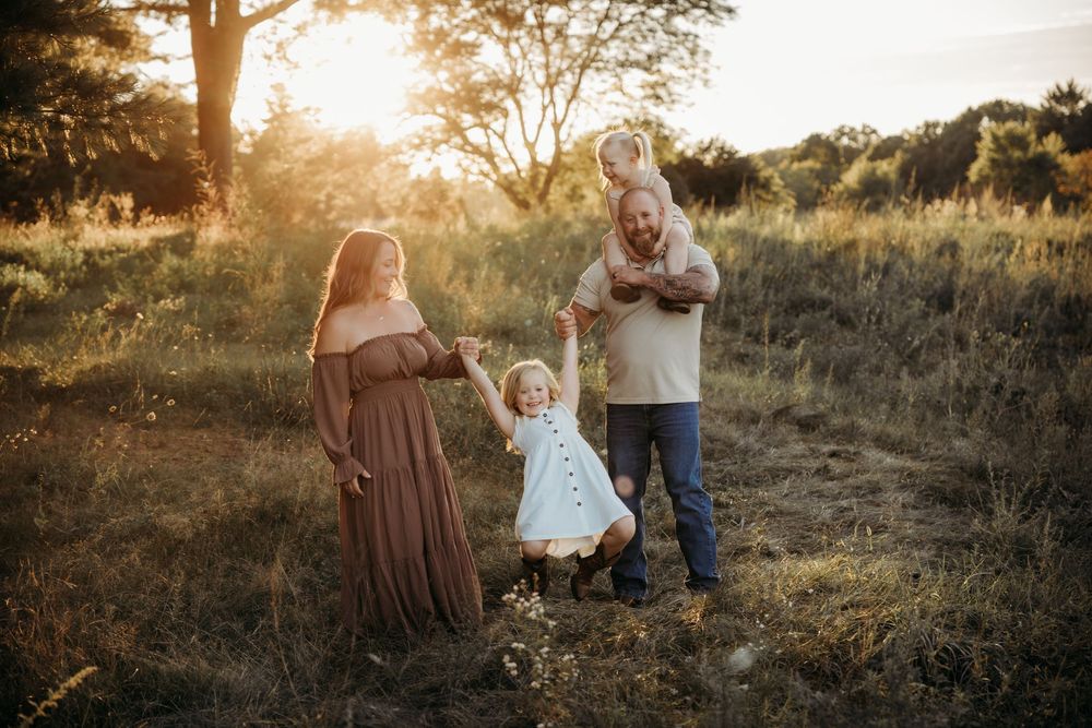 Family maternity photo at golden hour in a field, parents swinging toddler girl in white dress.