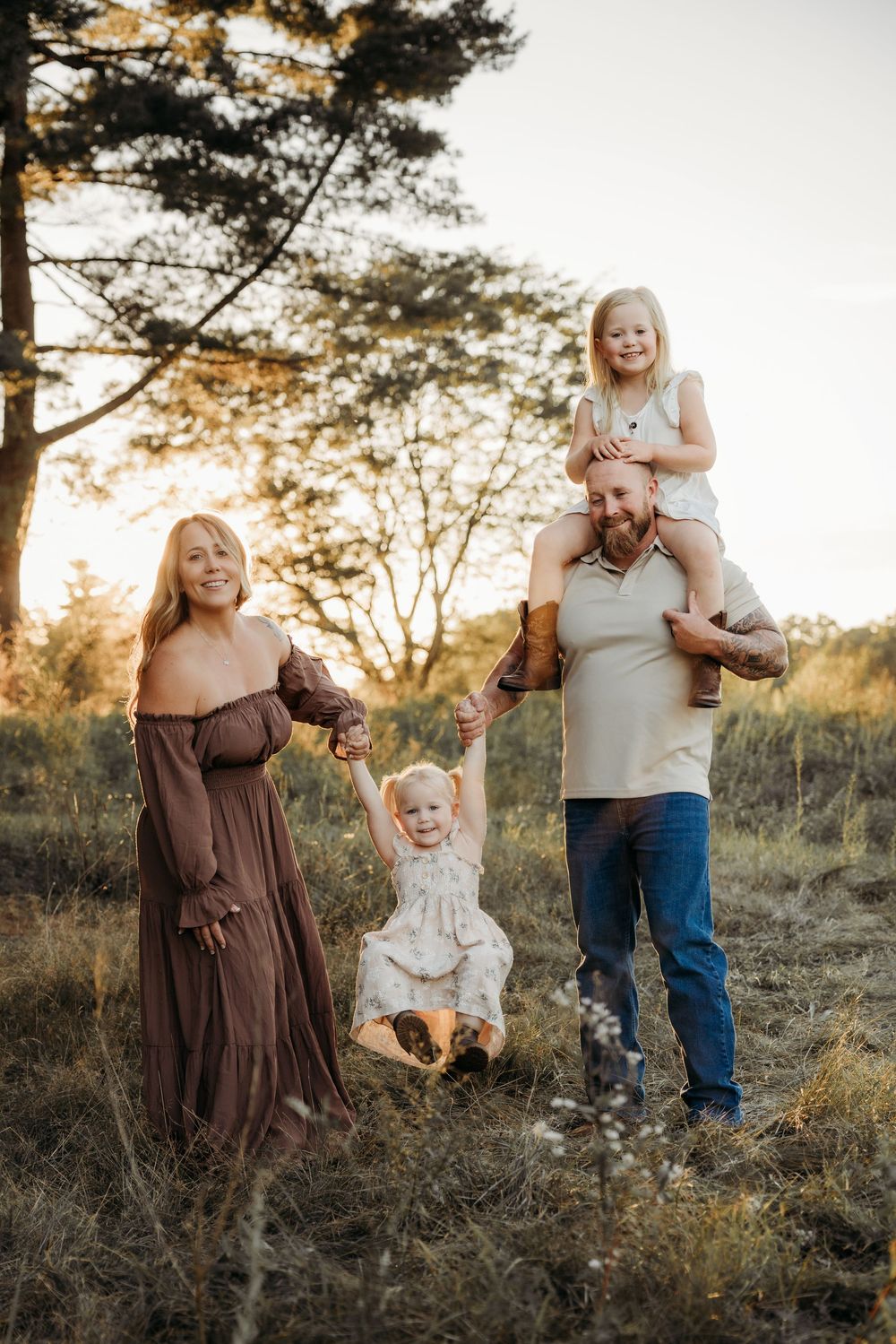 Family of four enjoying a golden sunset outdoors, mom in brown dress, dad carrying girl on shoulders.