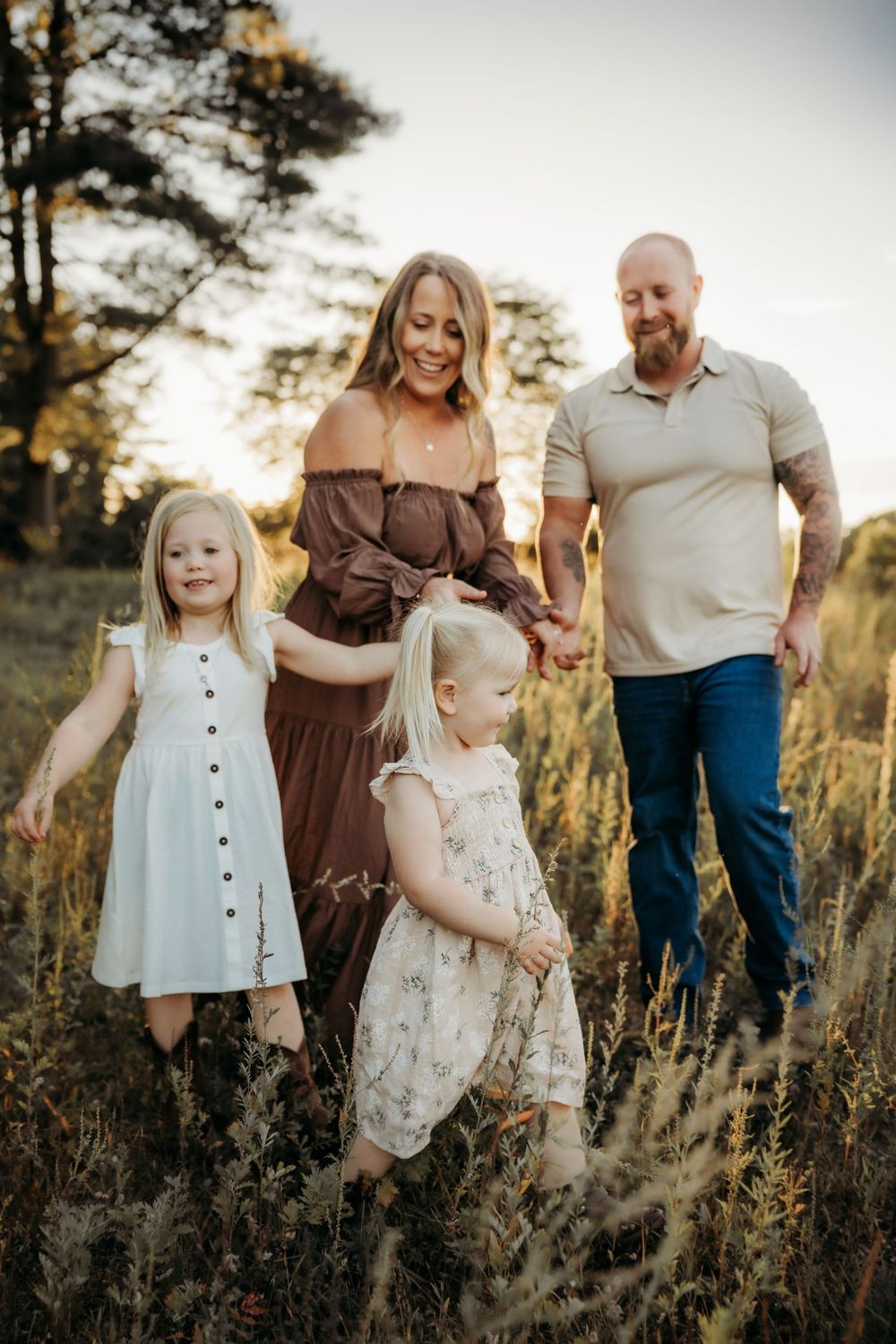Family of four posing in golden field at sunset, parents smiling with two young blonde girls in summer dresses.