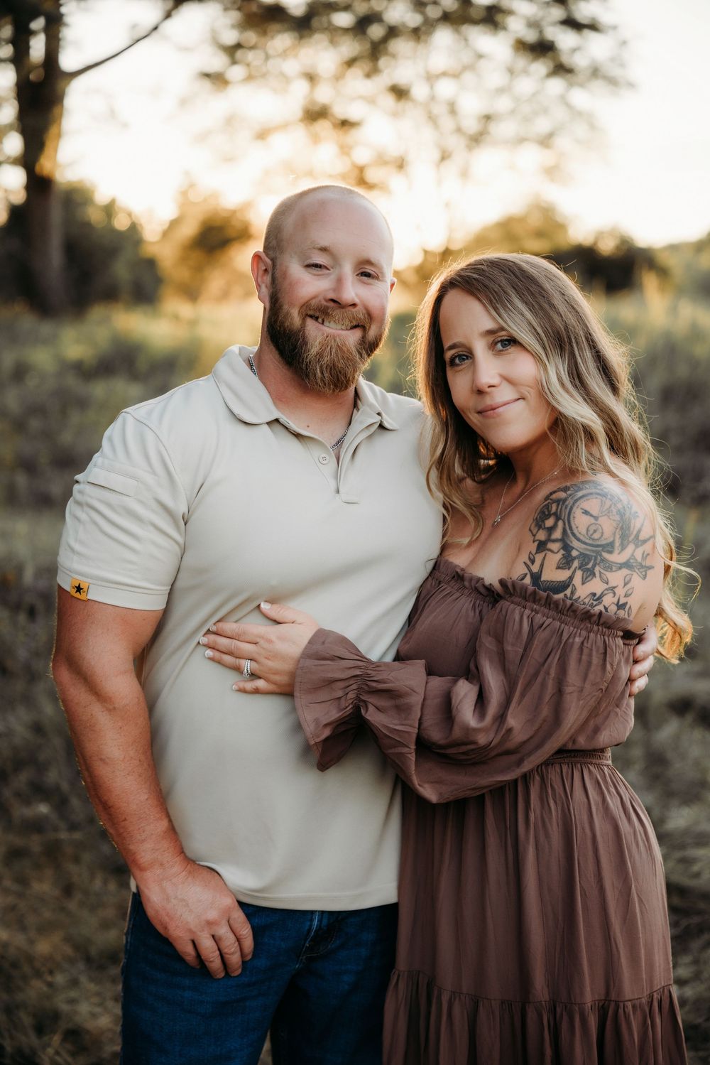 A smiling bald man with a beard and a woman with tattoos in a mauve dress embrace outdoors during golden hour.