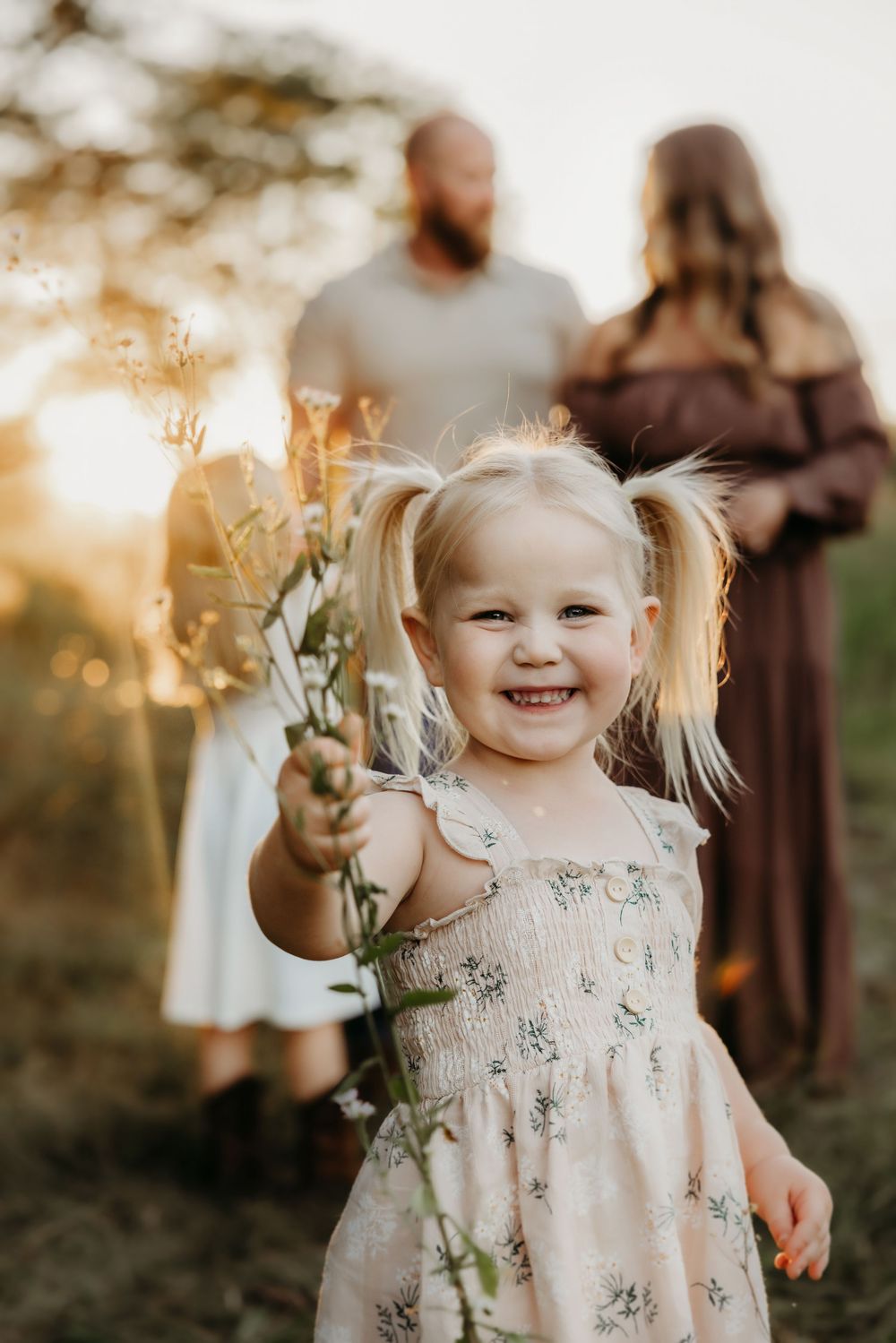 Smiling toddler girl with pigtails holding wildflowers outdoors at golden hour with parents in background.