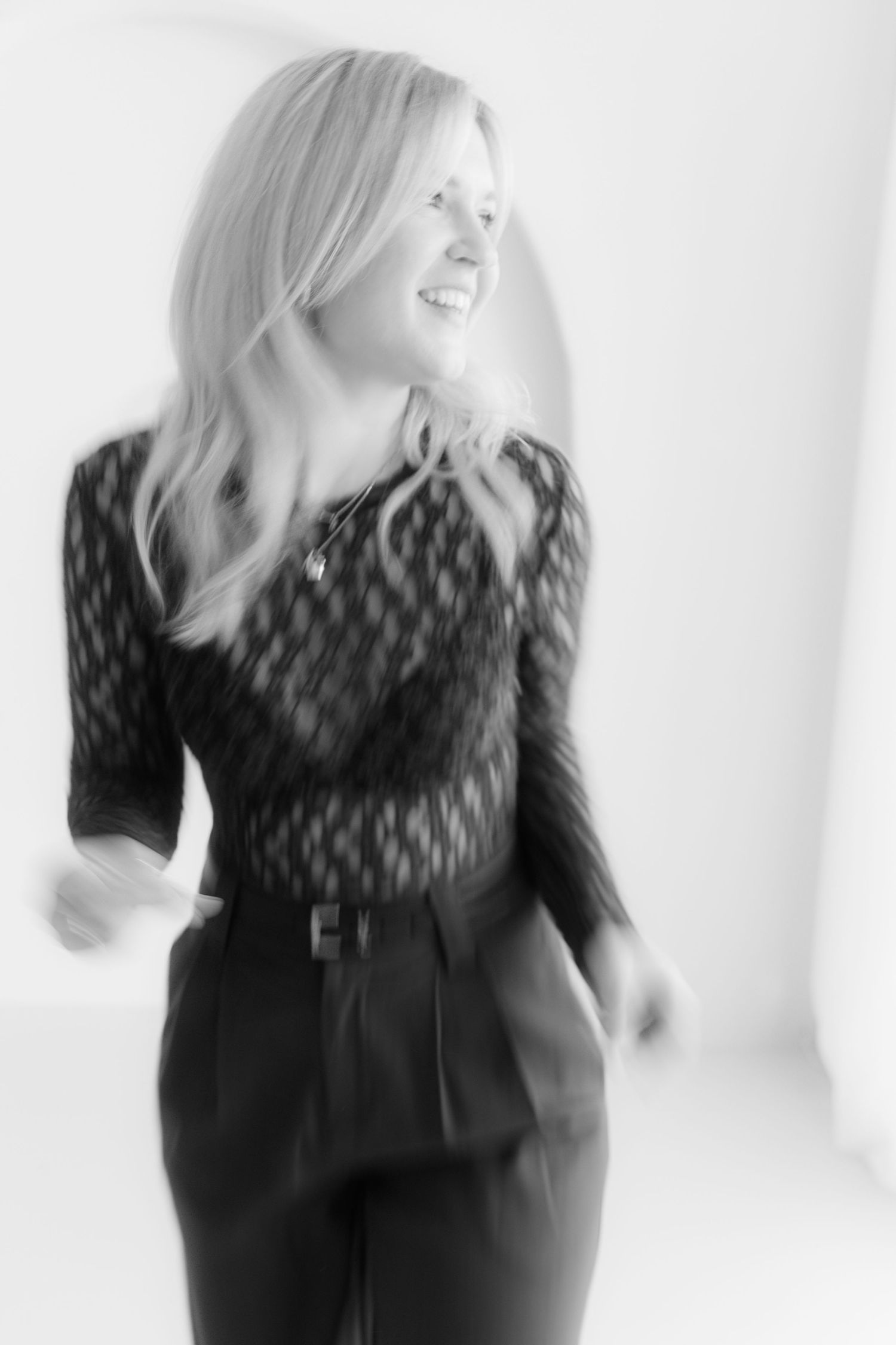 Black and white photo of a smiling blonde woman in a patterned top and belted trousers, captured mid-movement.