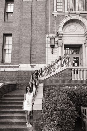 Group of women in white dresses and sashes posing on steps of a brick building in black and white photo.