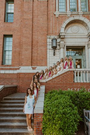 Group of young women in white dresses and sashes posing on steps of a brick university building.