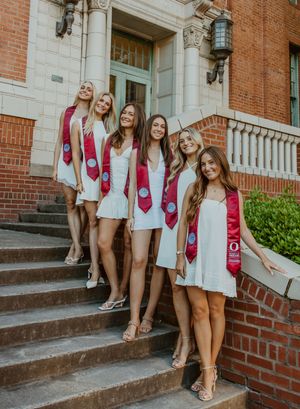 Five female graduates in white dresses with red graduation stoles on university steps.