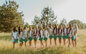 Group of young women in matching green skirts and white tops posing outdoors in a sunny field with trees.