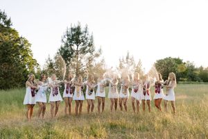 Bridesmaids in white dresses toss bouquets in a sunlit field, celebrating together in a joyful outdoor wedding photo.