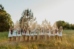 Group of women in white dresses tossing confetti in a sunny meadow with trees in the background.