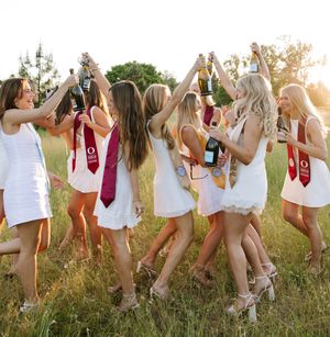 Group of young women in white dresses celebrating outdoors, raising champagne bottles in a sunny field.