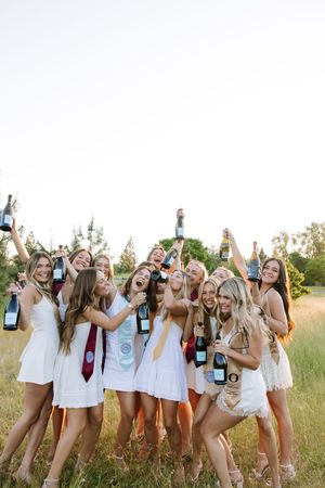 Group of young women in white dresses celebrating outdoors holding champagne bottles in a sunny field.