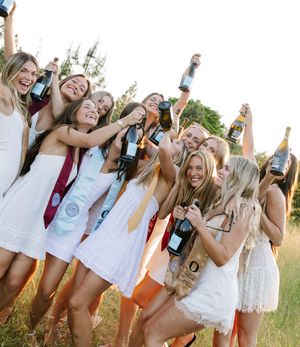 Smiling women in white dresses toasting with wine bottles during an outdoor graduation celebration.