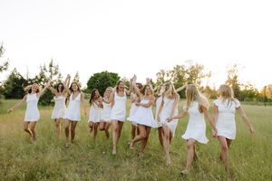 Group of women in white dresses dancing joyfully in a sunlit green meadow at golden hour.