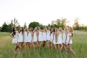 Group of women in white dresses posing together in a sunny green meadow.