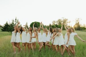 Group of young women in white dresses posing joyfully in a green meadow at golden hour.