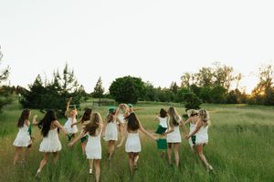 Group of girls in white dresses holding hands in a circle in a green field at sunset.