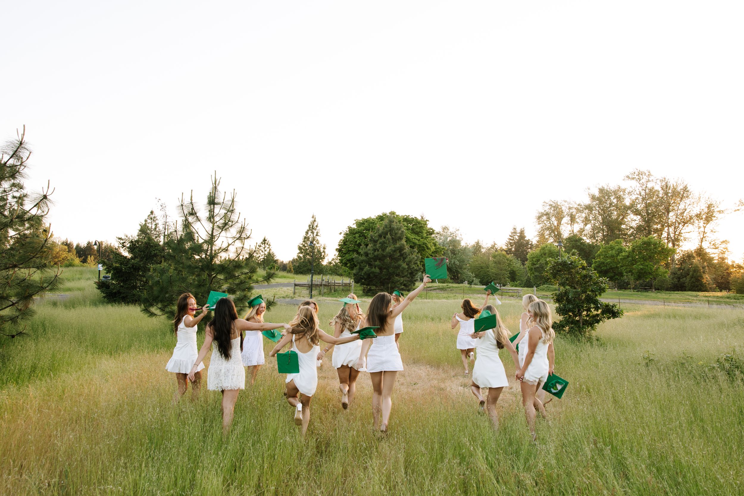 Group of girls in white dresses celebrating outdoors in a green meadow with trees in the background.