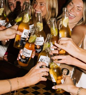 Group of young women laughing and clinking Corona Extra beer bottles with lime wedges at a bar celebration.