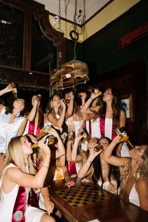 Group of young women in white dresses drinking beer bottles at a bar celebration event.