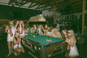 Group of young women in white dresses gathered around a pool table in a bar, smiling and posing together.