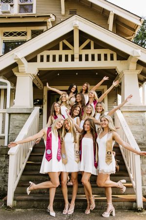 Group of young women in white dresses with graduation sashes posing on house steps in a celebratory photo.