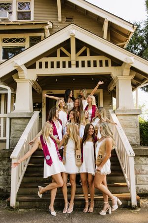 Group of young women in white dresses and sashes posing on steps of a house, celebrating graduation.