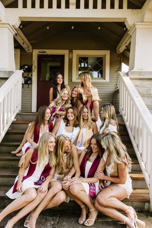 Group of smiling women in maroon sashes and white outfits sitting on porch steps together.