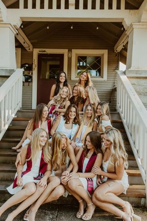 Group of young women in white and maroon outfits posing together on porch steps of a house.