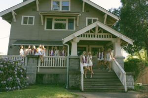 Group of young women in white outfits posing on the porch and steps of a green craftsman-style house.