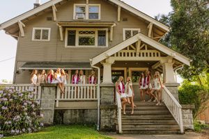 Group of women in white dresses with colorful sashes posing on porch of craftsman style house.