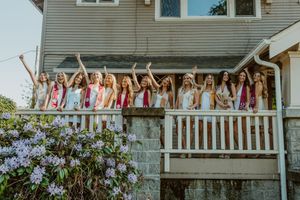 Group of young women in white dresses with sashes celebrating on a porch, waving and cheering together.