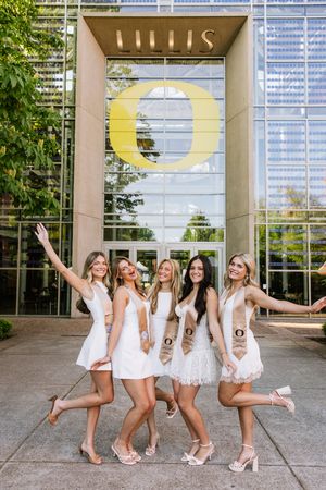 Five women in white dresses with graduation stoles posing at University of Oregon.