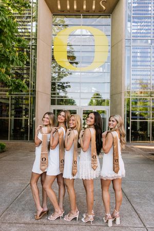 Five women in white dresses with UO graduation stoles posing at University of Oregon.