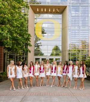 Group of female graduates in white dresses with colorful sashes posing outside Lillis building at University of Oregon.