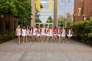 Large group of women in white dresses with maroon graduation stoles posing outside a modern building.