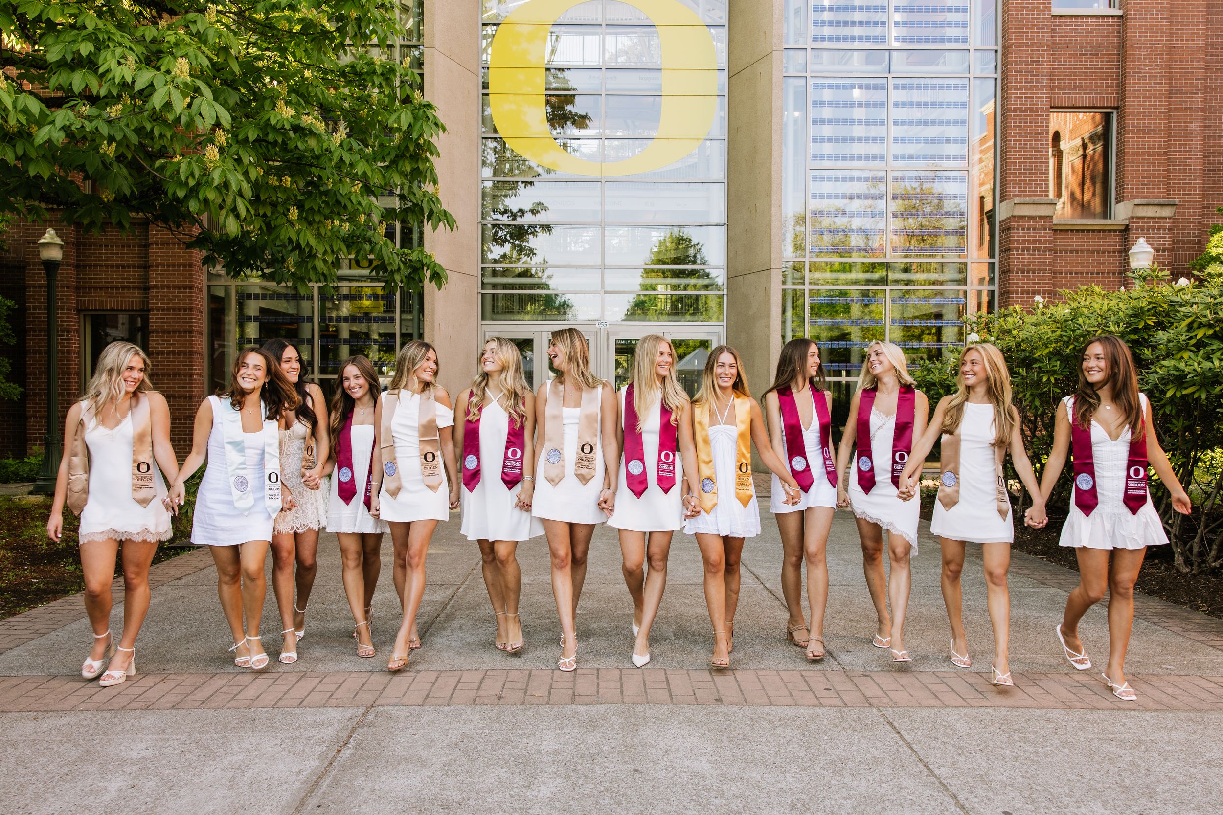 Women in white dresses and maroon graduation stoles holding hands in a line outside a university building.