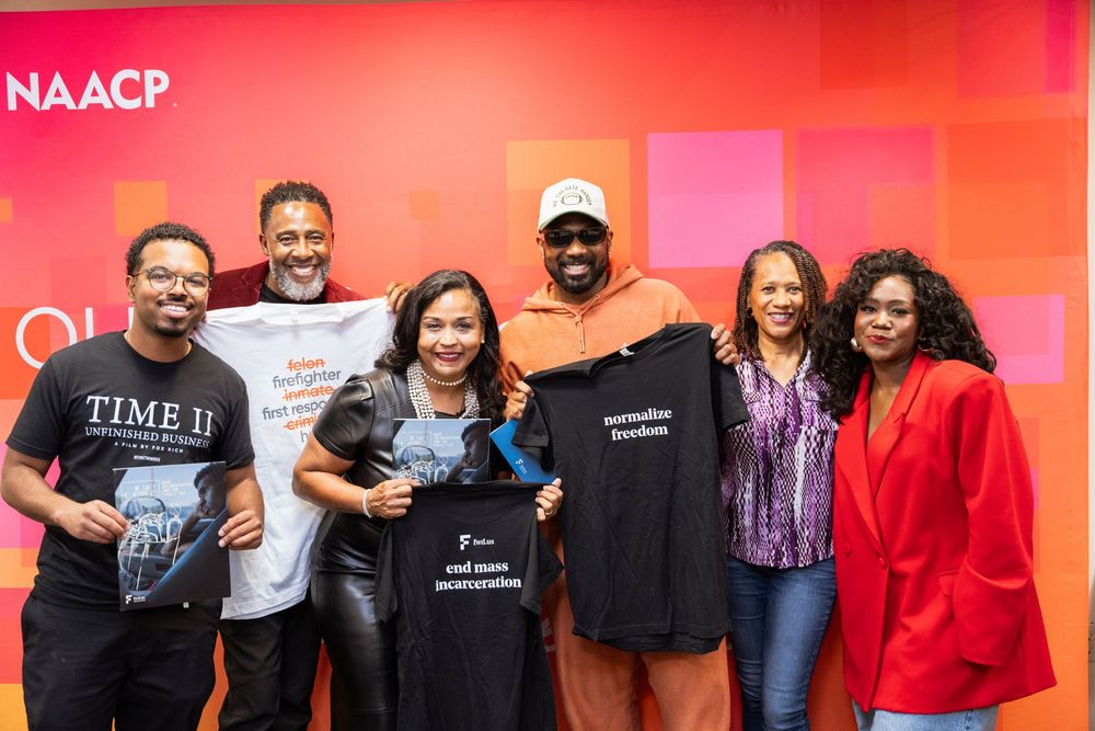 Six people posing together in front of an NAACP backdrop, some holding t-shirts at a community event.