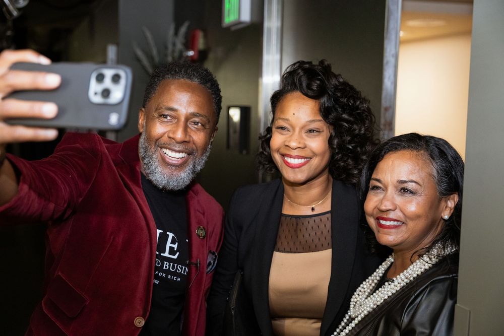 Three people smile together for a selfie at an indoor event, with a man in a red velvet blazer holding the phone.