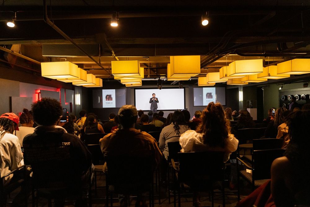 Audience watching a presenter on stage in a dimly lit conference room with yellow pendant lights overhead.