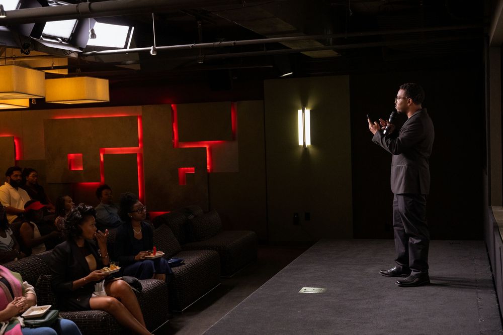 A speaker presents to an audience in a dimly lit venue with red geometric wall decor.