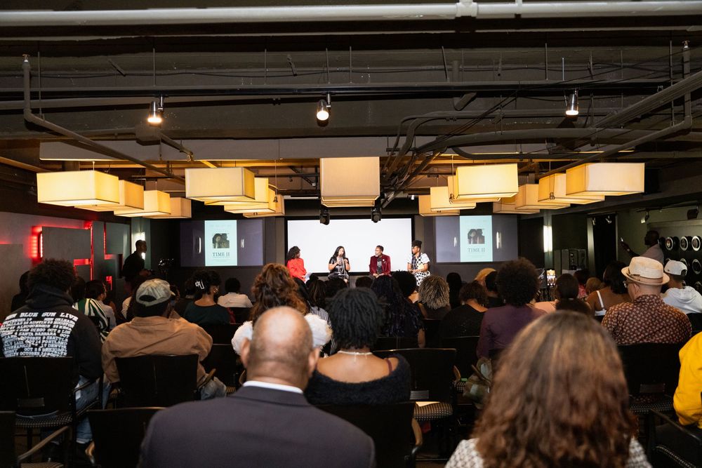 Panel discussion event in a modern venue with square pendant lights, audience seated facing three speakers on a stage.