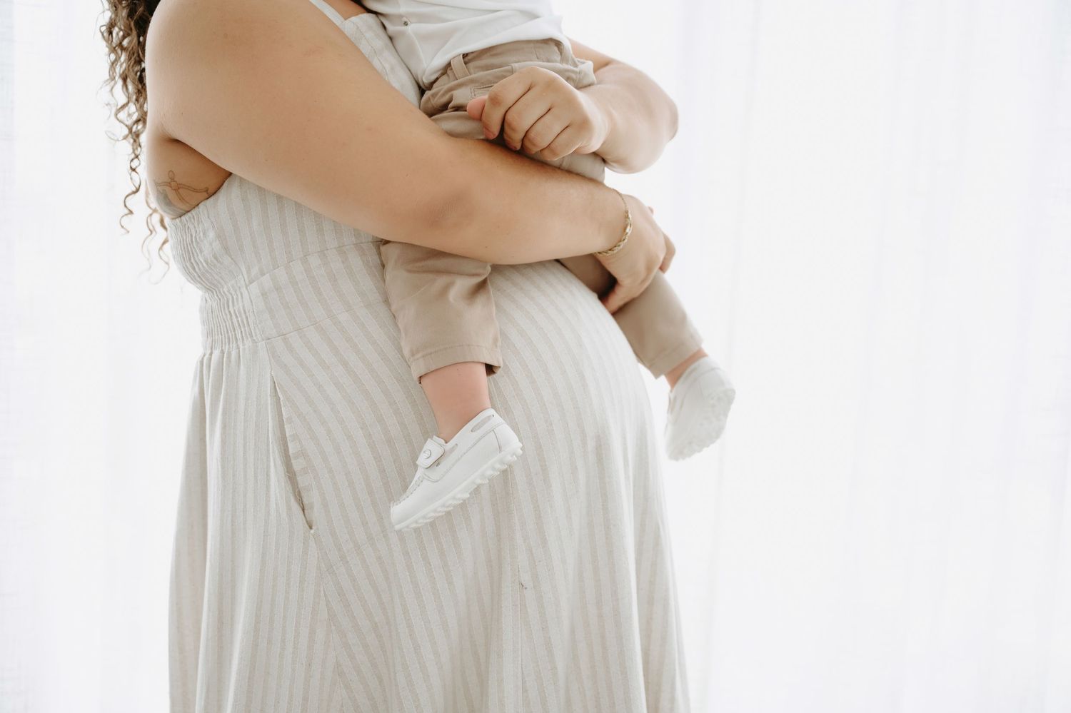 A mother in a striped dress holds her baby against her pregnant belly near a bright window.