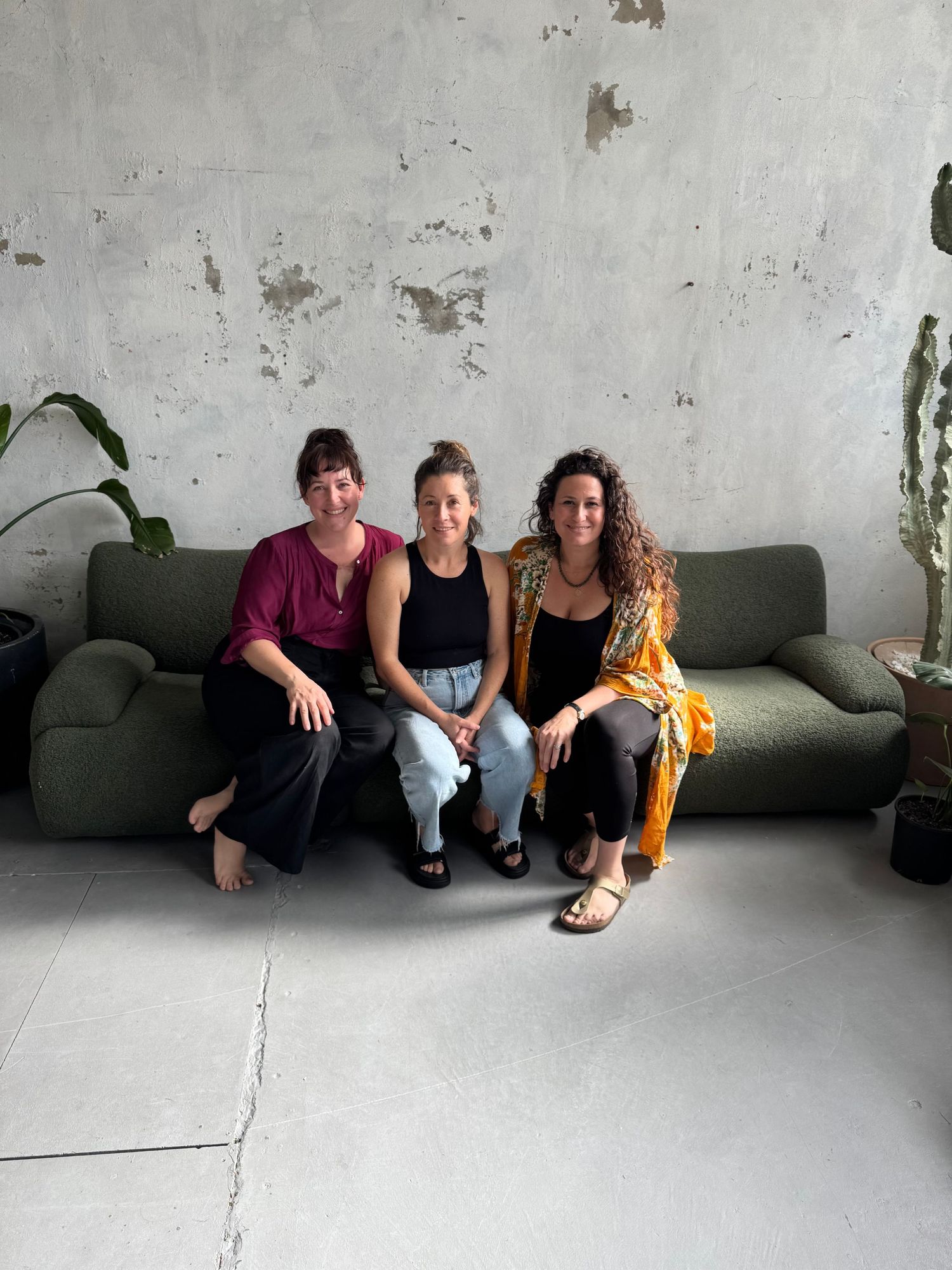 Three women sitting together on a green couch against a textured gray wall with plants nearby.