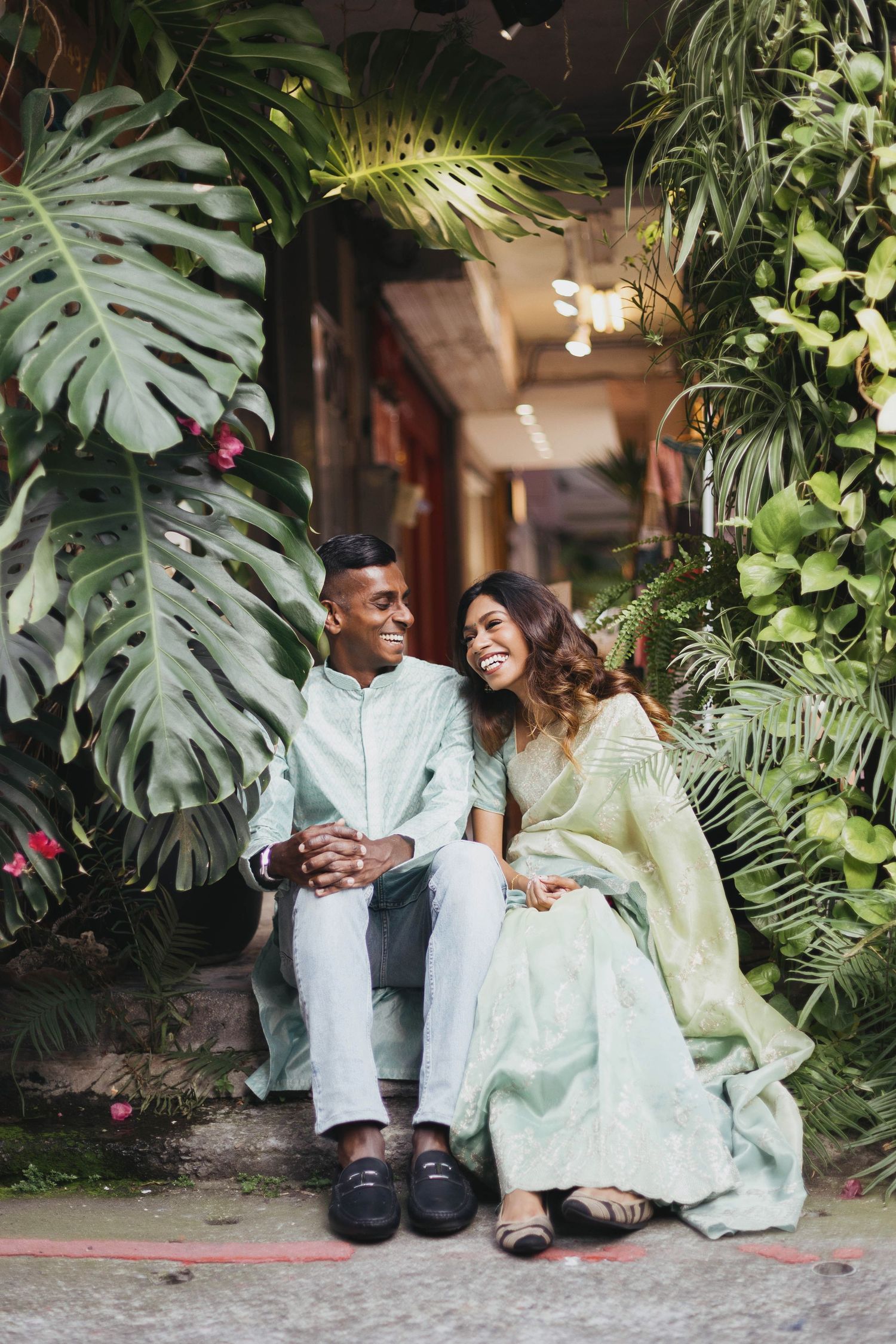 A smiling couple in white outfits sitting on vintage shop steps surrounded by plants and old utility meters.