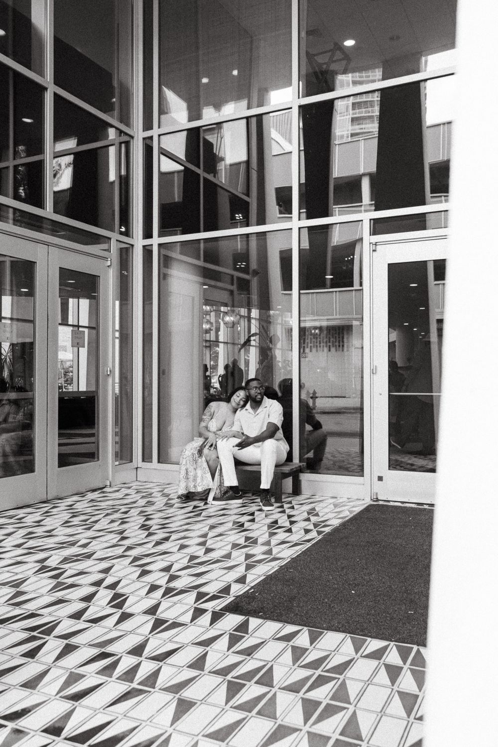 Couple sitting together on a bench outside a modern building with geometric tiled floors, black and white photo.