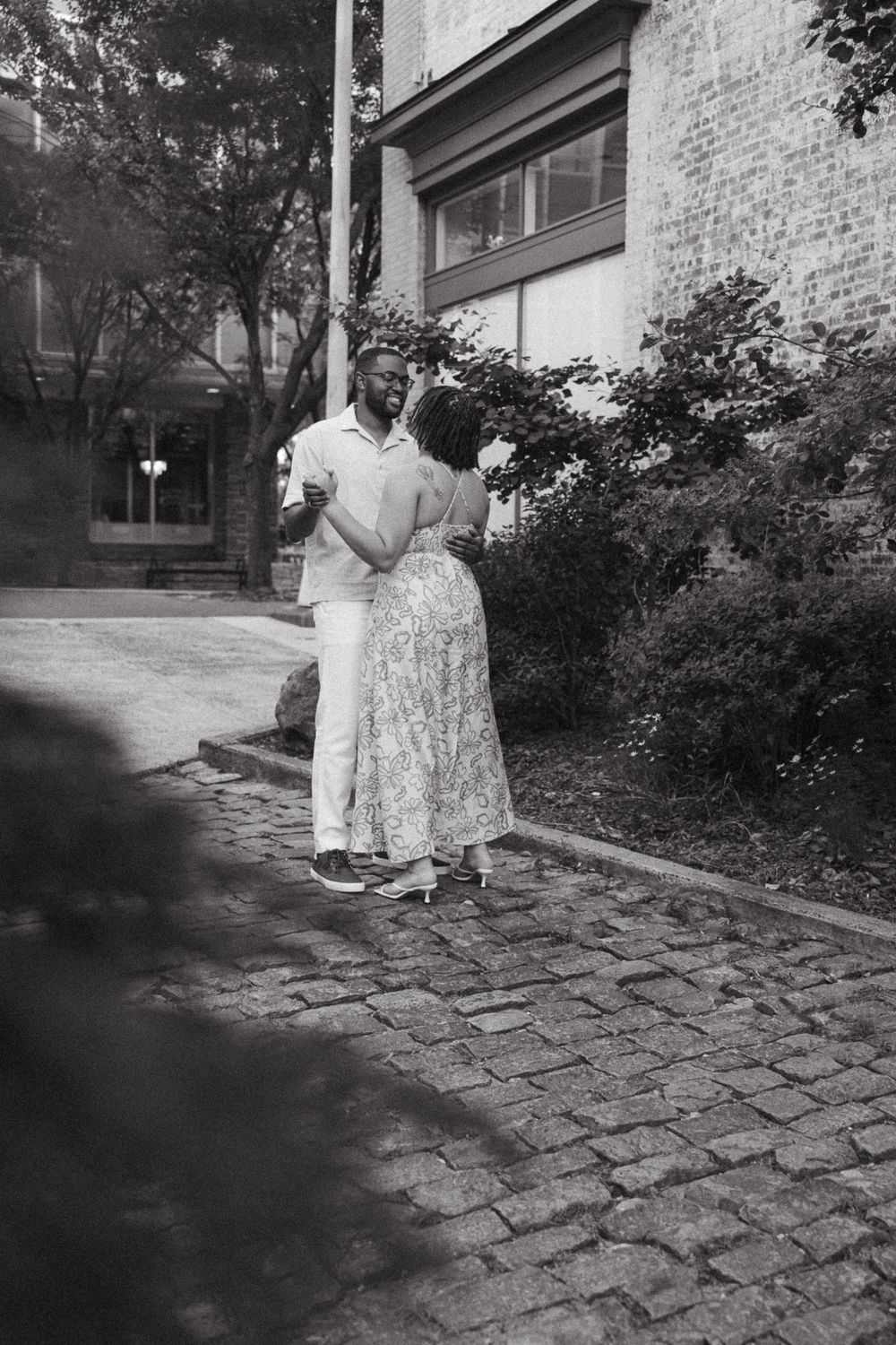 Black and white photo of a couple embracing on a cobblestone street beside a brick building and lush greenery.