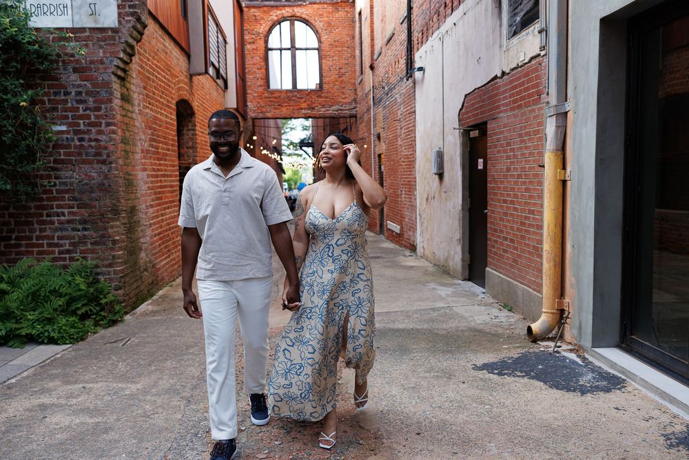 Couple walking hand-in-hand through a charming brick alleyway, man in white outfit and woman in floral dress.