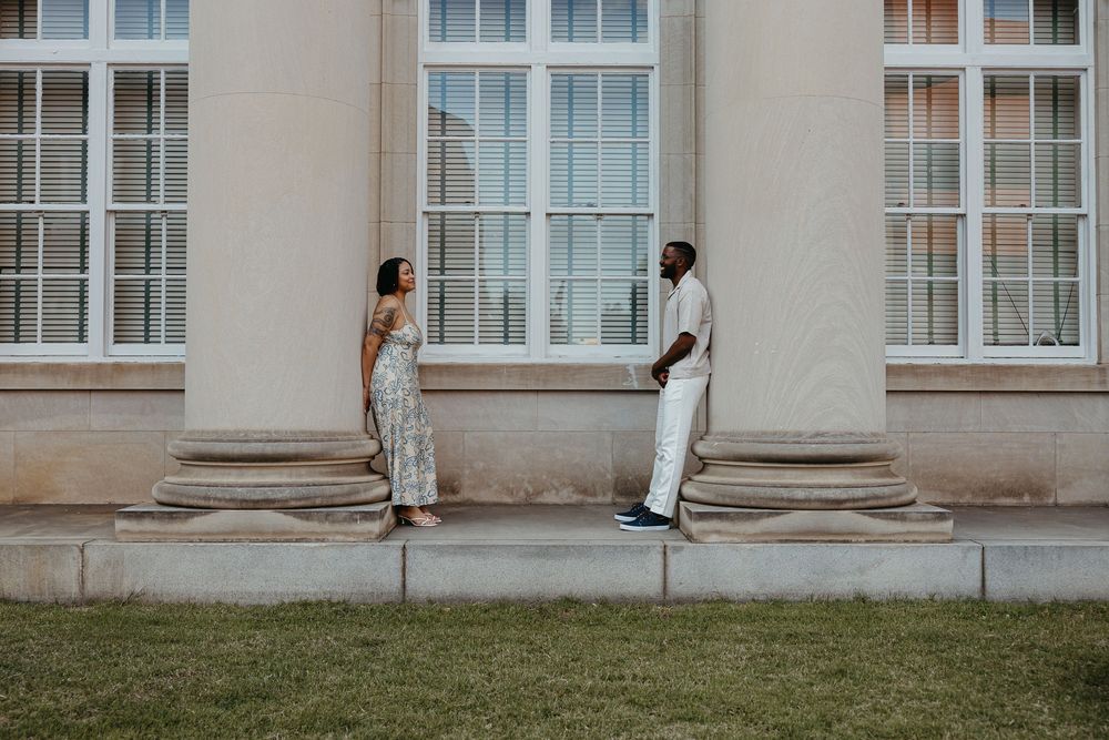 A couple stands apart by elegant columns of a classical building, dressed in white and floral outfits on a sunny day.