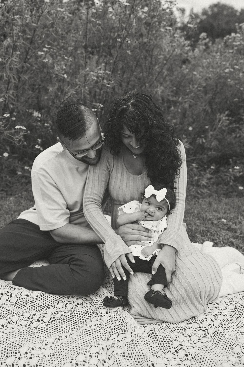 Black and white photo of parents sitting on lace blanket outdoors, lovingly gazing at their newborn baby.